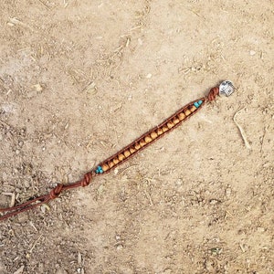 以下が含まれることがあります： 茶色の革のブレスレットにターコイズ色のビーズと銀のチャームが付いています。ブレスレットは結び目で結ばれており、茶色の土の表面に置かれています。