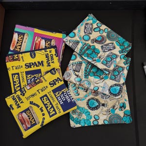 May include: A collection of fabric pouches with various designs. Some pouches feature a yellow background with the word "SPAM" and "Iconic Taste" printed on them. Other pouches have a beige background with turquoise and silver jewelry patterns.