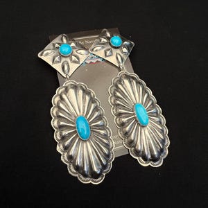 May include: Silver earrings with turquoise stones. The earrings feature a diamond-shaped top with a small turquoise stone and an oval-shaped bottom with a central turquoise stone. The design includes a sunburst pattern.