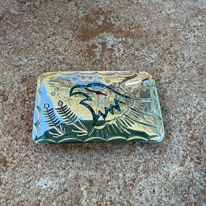 May include: A rectangular silver belt buckle with an engraved eagle design. The eagle's head and wing feathers are detailed with turquoise accents. The buckle has a decorative border and is likely made of metal.