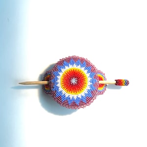 May include: A beaded hair stick with a colorful circular design in shades of red, orange, yellow, white, and blue. The stick is made of wood and has a small bead at the end.