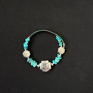May include: A bracelet featuring turquoise chip beads, three square silver-toned metal charms with embossed geometric patterns, and a thin band of iridescent multi-colored seed beads. The components are arranged in a circular pattern.