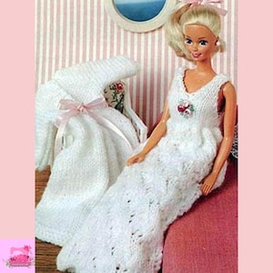 May include: A doll wearing a white knitted gown with a V-neck and floral embellishment. A matching white knitted wrap with a pink ribbon is nearby. The doll is seated on a pink surface, with a striped background.