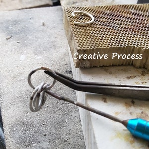 May include: A silver ring being worked on with a pair of silver tweezers. The ring is being held in place by the tweezers and a small, blue, metal tool. The background is a light grey surface with the words 'Creative Process' printed on a brown surface.