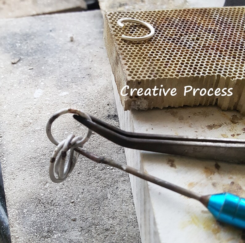 May include: A silver ring being worked on with a pair of silver tweezers. The ring is being held in place by the tweezers and a small, blue, metal tool. The background is a light grey surface with the words 'Creative Process' printed on a brown surface.