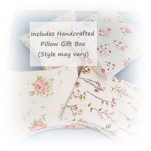 May include: A set of four handcrafted pillow gift boxes with floral patterns. The boxes are made of white cardstock with pink and green floral designs. The text "Includes Handcrafted Pillow Gift Box (Style may vary)" is printed on one of the boxes.