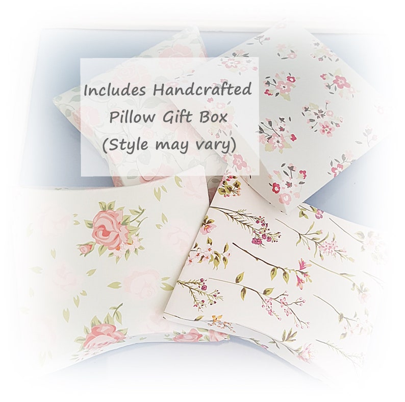 May include: A set of four handcrafted pillow gift boxes with floral patterns. The boxes are made of white cardstock with pink and green floral designs. The text "Includes Handcrafted Pillow Gift Box (Style may vary)" is printed on one of the boxes.