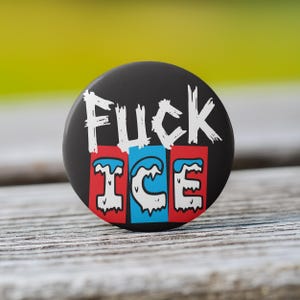 May include: A black button with the words "FUCK ICE" in white, stylized lettering. Below, the word "ICE" is in a red, blue, and white design. The button is round and sits on a wooden surface.