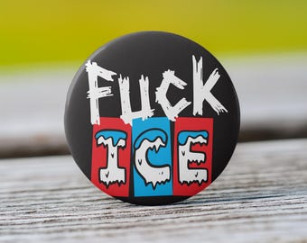 May include: A black button with the words "FUCK ICE" in white, stylized lettering. Below, the word "ICE" is in a red, blue, and white design. The button is round and sits on a wooden surface.