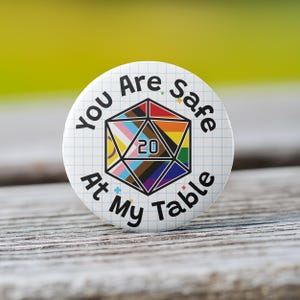 May include: A white button with a grid pattern and the text "You Are Safe At My Table" in black. A 20-sided die with a rainbow design is in the center of the button.