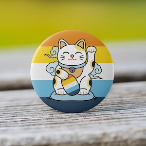 May include: A round button featuring a cartoon Maneki-neko cat with a raised paw. The cat is white with black spots and a blue collar, set against a striped background of orange, yellow, and blue. The button is on a wooden surface.