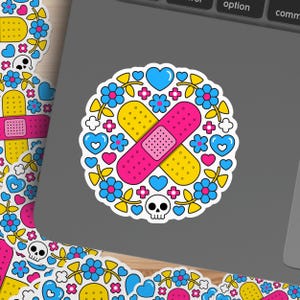 May include: A circular sticker featuring a pink and yellow bandage design, surrounded by blue flowers, hearts, and a small skull. The sticker has a white border and is placed on a gray surface.