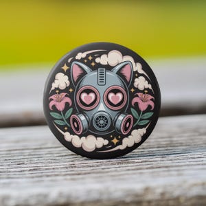 May include: A round, black button featuring a cat-like gas mask design with pink heart-shaped lenses and ears. Pink lilies and white clouds surround the mask. The button is set against a blurred green and wooden background.