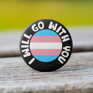 May include: Black button with the text "I will go with you" in white around a transgender pride flag.