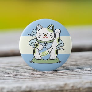 May include: A round button featuring a cartoon Maneki-neko cat with a raised paw. The cat is white with black spots and green accents, set against a light blue background with a cream stripe. The cat sits on a green cushion.