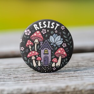 May include: A black button with the word "RESIST" in white lettering. The button features a whimsical illustration of a purple house with a door and window, surrounded by red and white mushrooms, flowers, and leaves on a black background.