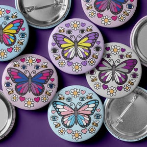 May include: A collection of colorful round pinback buttons with butterfly designs. Each button features a different butterfly with various colors, surrounded by flowers, hearts, eyes, and bees. The buttons are on a purple background.