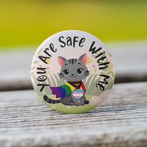 May include: A round button with a gray cat wearing a rainbow cape and a transgender pride heart. The text on the button reads "You Are Safe With Me."