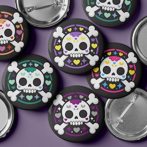 May include: A collection of black, round buttons featuring a sugar skull design. Each button has a white skull with crossbones, surrounded by colorful hearts, stars, and a decorative border. The buttons are scattered on a purple surface.