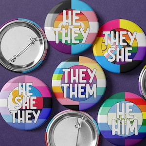 May include: A set of eight colorful button pins with different pride flag designs and text including "He They", "They She", "They Them", "He She They", and "He Him".