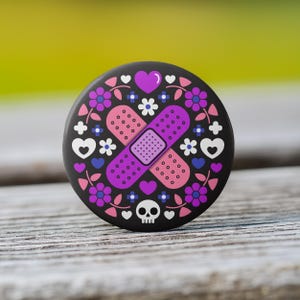 May include: A round, black button with a colorful design. The design features a purple bandage, pink and purple flowers, white hearts, and a small skull. The button is set against a blurred green and wood background.