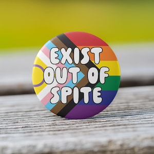 May include: A round button with the text "EXIST OUT OF SPITE" in white, set against a rainbow background. The button features various pride flag designs, including the classic rainbow, transgender, and progress pride flags.