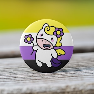 May include: A round button with a non-binary pride flag design. The button features a cartoon unicorn character holding a flower. The unicorn has a white body, yellow hair, and a purple flower. The background is a blurred wooden surface.