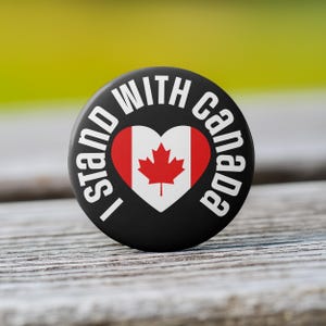 May include: Black button with white text that reads "I stand with Canada" and a red maple leaf inside a white heart.