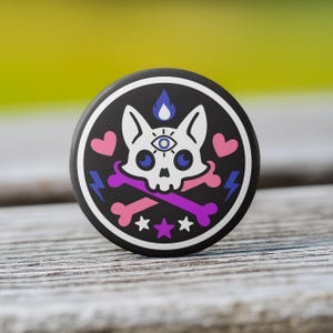 May include: A round, black button with a white border featuring a skull with an eye in the forehead, crossed bones, hearts, stars, and lightning bolts. The design incorporates pink, purple, blue, and white elements.