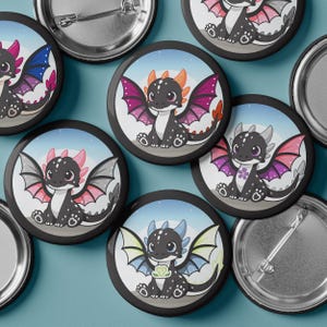 May include: A collection of black button pins featuring cartoon dragons with various wing colors, including pink, purple, and blue. Each dragon has a unique design, set against a light blue and white background. The pins are round with a metal backing.