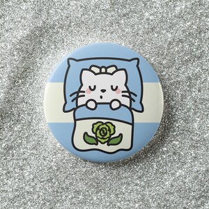 May include: A round, blue button featuring a cartoon cat sleeping in a bed. The cat is white with closed eyes and a small bow. The bed has a blue and white striped design with a green rose on the blanket.