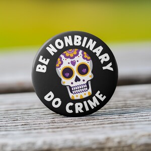 May include: Black button with white text that reads "Be Nonbinary Do Crime". A white sugar skull with purple and yellow details is in the center.