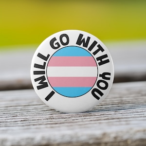 May include: White button with black text that reads "I will go with you" and a transgender pride flag in the center.
