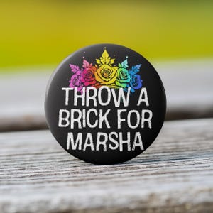 May include: A black button with a rainbow crown of roses and the text "THROW A BRICK FOR MARSHA" in white.