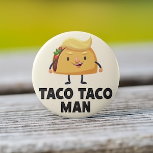 May include: A round, off-white button with a cartoon taco character wearing a blonde wig. The taco has arms, legs, and a smiling face. The text "TACO TACO MAN" is printed in bold, black letters below the character.