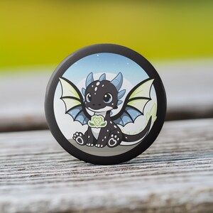 May include: A round, black button featuring a cartoon dragon with black scales, blue horns, and light green wings. The dragon is holding a green flower. The background is a light blue sky with white stars.
