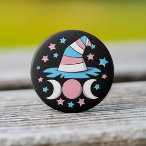 May include: A round black button featuring a witch hat in the colors of the transgender flag, surrounded by stars and crescent moons. The hat is blue, white, and pink. The button is on a wooden surface.