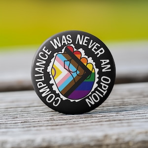 May include: Black button with white text that reads "Compliance was never an option". A raised fist with a rainbow, transgender, and black and brown pride flag design is in the center of the button.