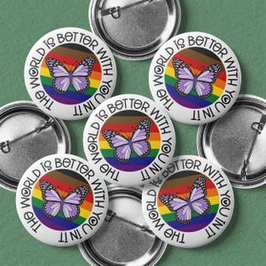 May include: A collection of white button pins with a rainbow and butterfly design. Each pin has the text "THE WORLD IS BETTER WITH YOU IN IT" around the edge. The butterfly is purple with black details.