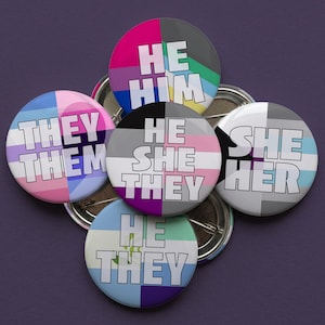 May include: Five circular pin-back buttons featuring various pronoun combinations in bold white text. The buttons display diverse color patterns including pink, blue, grey, green, and purple, with text reading HE HIM, THEY THEM, HE SHE THEY, SHE HER, and HE THEY.