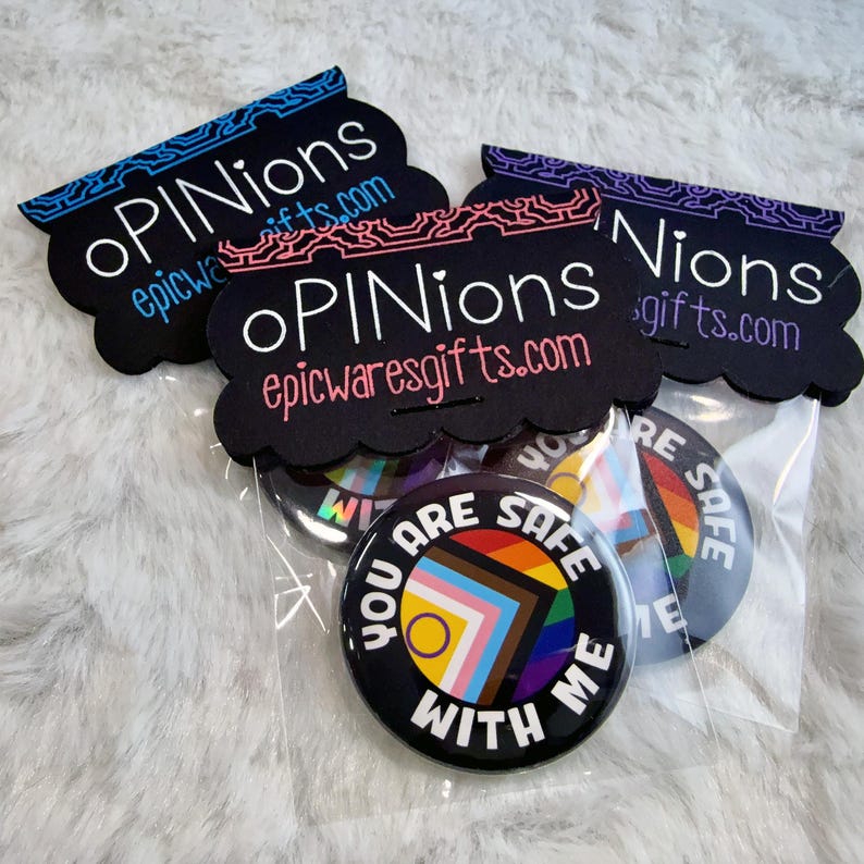 K&ouml;nnte beinhalten: Eine Sammlung schwarzer Ansteckbuttons mit dem Text "YOU ARE SAFE WITH ME" und einem farbenfrohen geometrischen Design. Die Buttons sind mit dekorativem schwarzen Karton mit dem Text "oPINions" und "epicwaresgifts.com" verpackt.