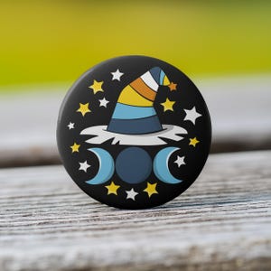 May include: A round black button with a colorful witch hat design. The hat is striped with yellow, orange, and blue. The button also features stars and crescent moons in yellow and white, creating a celestial theme.