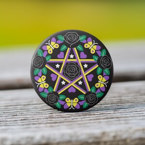 May include: A black button with a pentagram design. The design includes a yellow and purple star, black roses, butterflies, and hearts. The button is set against a blurred green and wood background.