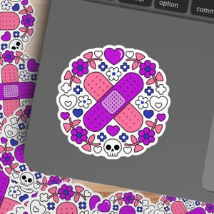 May include: A circular sticker featuring a purple and pink bandage design, surrounded by flowers, hearts, and a skull. The sticker has a white border and is placed on a gray surface.