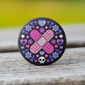 May include: A black button pin featuring a pink bandage design with a white dotted pattern, crossed over each other. The pin is decorated with blue and purple flowers, hearts, and a small skull.
