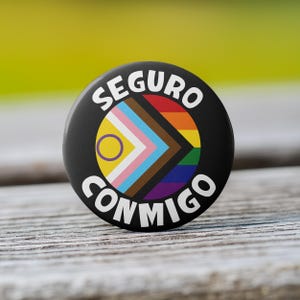 May include: A black button with white text that reads "Seguro Conmigo" and a rainbow and gender-inclusive pride flag design.