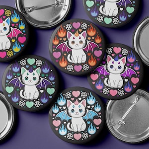 May include: A collection of black button pins featuring a white cat with bat wings, a horn, and a pentagram. The cat is surrounded by hearts and flames in various colors. The pins are on a purple background.