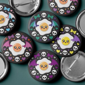 May include: A collection of black round buttons featuring a cartoon fried egg with bat wings, skulls, and hearts. The egg has a smiling face and is surrounded by bows, skulls, and hearts in various colors. The buttons are made of metal.