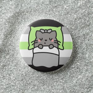 May include: A round button featuring a sleeping gray cat illustration. The cat is nestled on a green pillow under a gray blanket. The button has a striped background in gray, white, and green, with a black border.