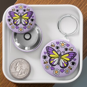K&ouml;nnte beinhalten: Ein Set aus zwei runden, lavendelfarbenen Kompaktspiegeln und einem Schl&uuml;sselanh&auml;nger. Jeder Spiegel zeigt ein Schmetterlingsdesign mit lila, gelben und wei&szlig;en Fl&uuml;geln, umgeben von Bienen, Blumen und Herzen. Eine M&uuml;nze liegt ebenfalls in der Schale.